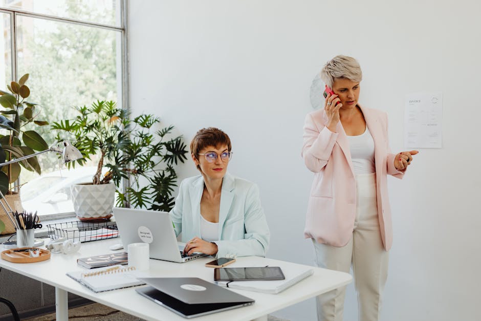 Two businesswomen working together in a modern office setting, using laptops and making phone calls.